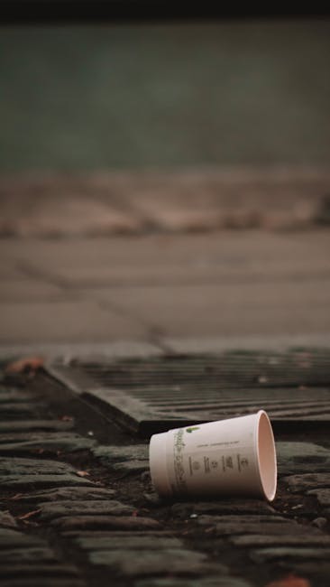 A discarded paper cup, lying on a cobbled pavement surface near a metal grate, with a blurred background of a wooden deck or platform and a patch of greenish grassy area. The paper cup appears to be m