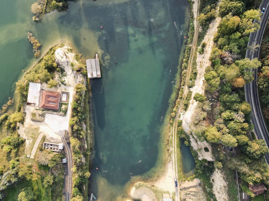 An aerial view of a narrow, elongated body of water surrounded by lush green trees and vegetation. On the left side of the water, there is a small building with a reddish-brown roof, adjacent to a doc