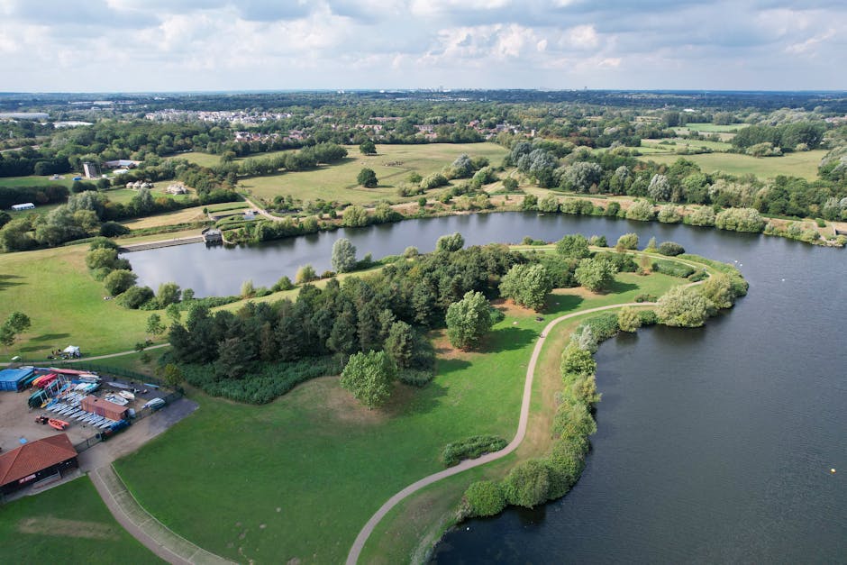 An aerial view of a narrow, elongated body of water surrounded by lush green trees and vegetation. On the left side of the water, there is a small building with a reddish-brown roof, adjacent to a doc
